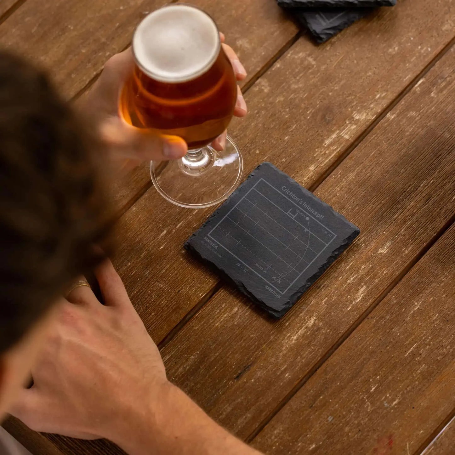 Person holding a glass of beer on a wooden table with a NRL play-book slate coaster.