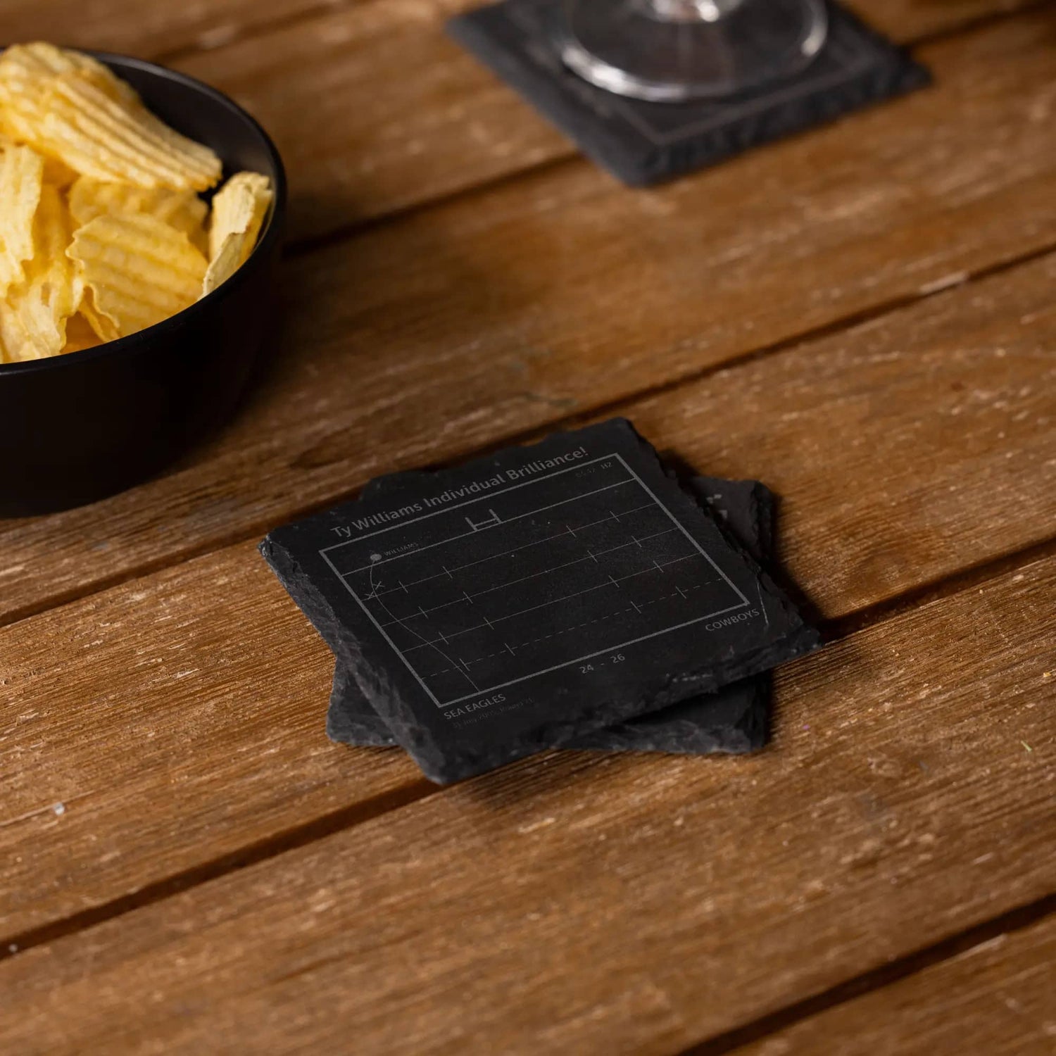 NRL playbook slate coasters  on a wooden surface next to a bowl of chips.